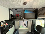 RV rental interior: overhead storage with an inverter, power outlets, and grey felt lining. A key panel and small window below.