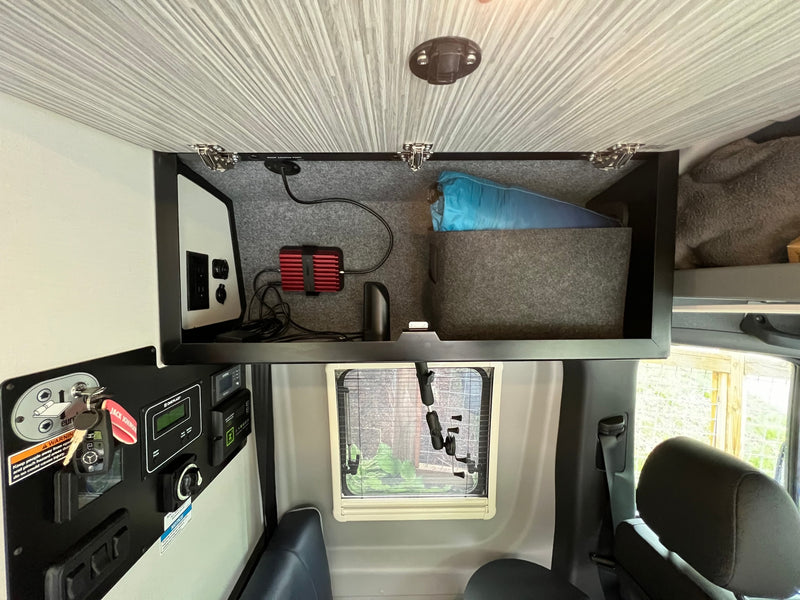 RV rental interior: overhead storage with an inverter, power outlets, and grey felt lining. A key panel and small window below. — photo 7
