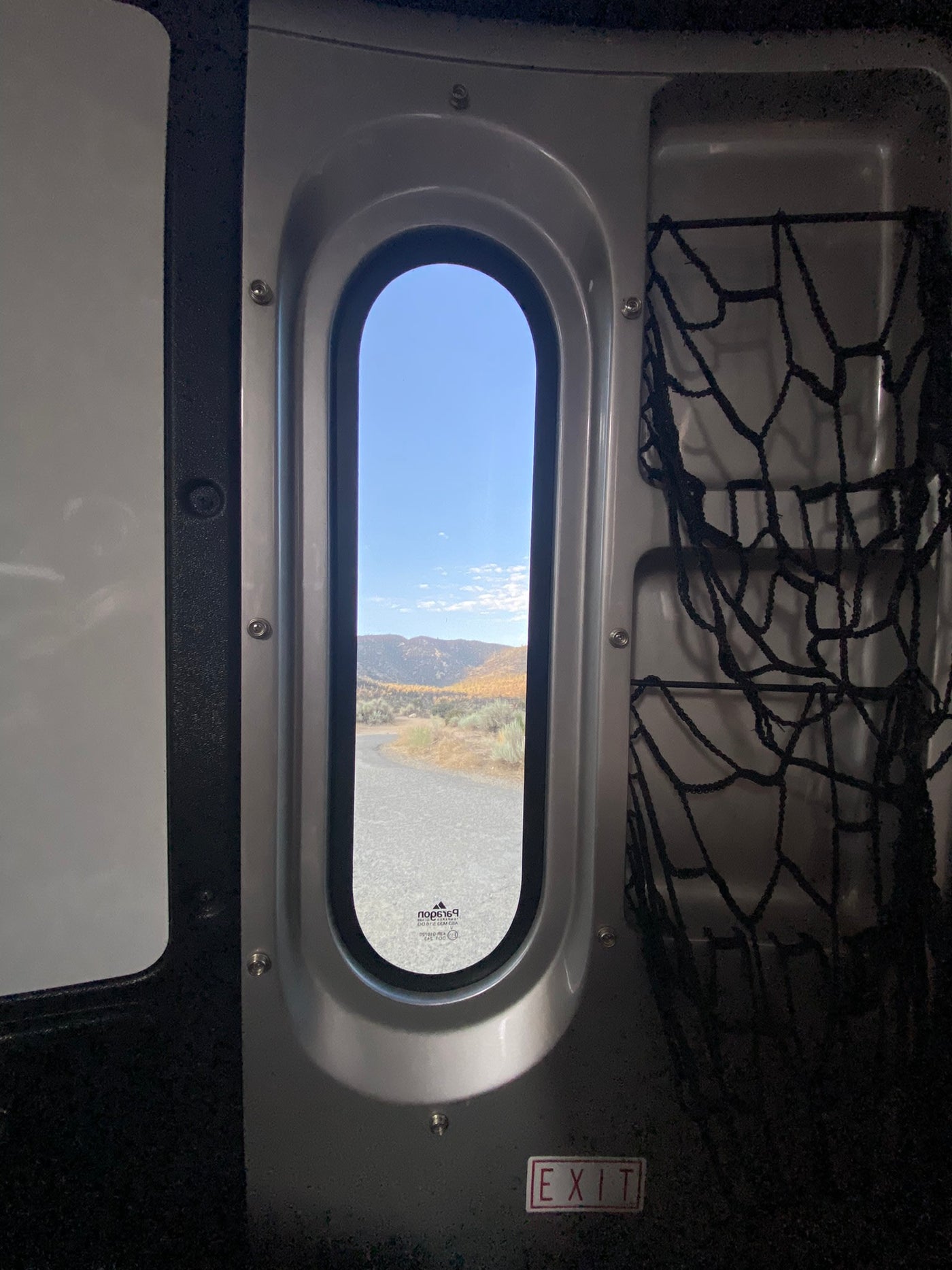 RV rental interior: oval window frames fall mountains, road. Storage nets & EXIT sign. — photo 15