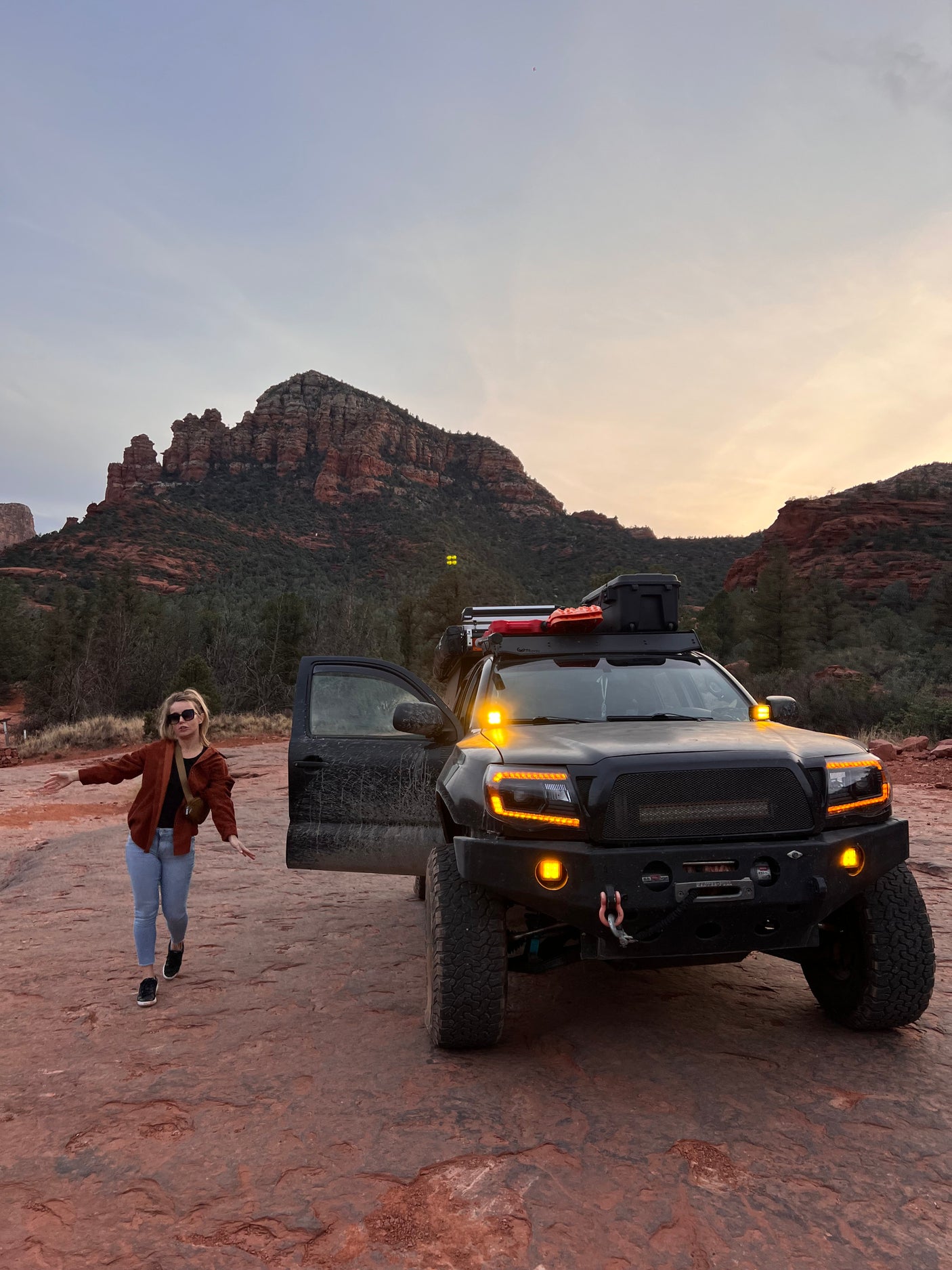 Woman poses by a custom off-road truck with bright lights in Sedona's red rock landscape, ideal for a rugged adventure or RV rental trip.