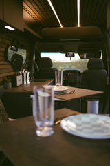 Cozy RV rental interior: wooden ceiling with lights, kitchenette with sink & speaker, dining table set for two, facing the driver's cabin.