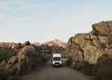 White RV rental van on a dusty road, flanked by boulders, with mountains glowing at sunset.