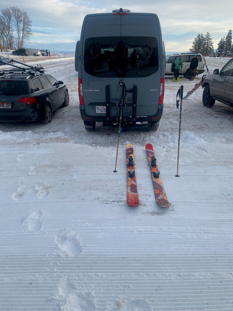 Skis and a pole rest on groomed snow beside a grey Mercedes Sprinter RV rental van, prepared for a winter day.
