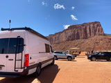 White RV rental parked on red dirt. Majestic red rock cliffs rise under a clear sky.