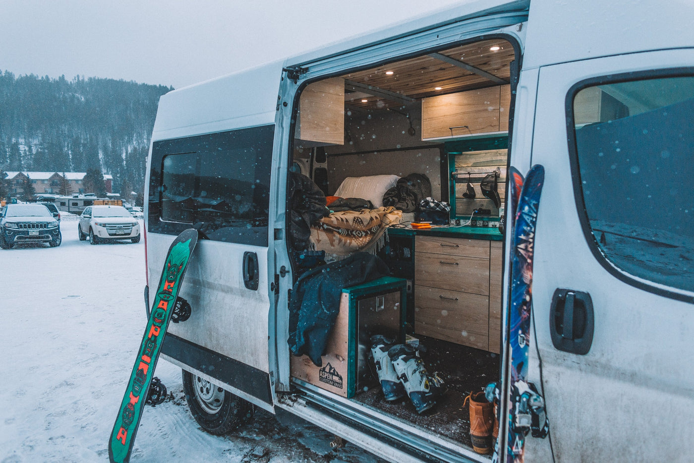Snowy RV rental van reveals cozy converted interior with bed, skis/snowboards. Ready for winter adventure. — photo 12