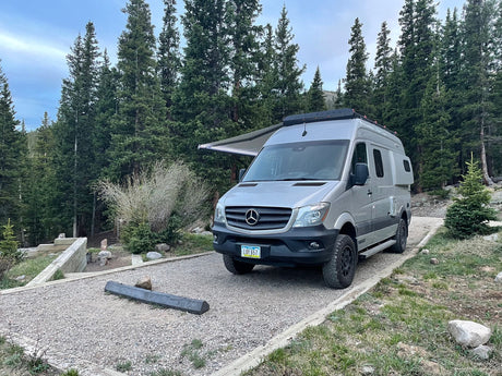 A silver Sprinter van rv rental is parked on a gravel campsite amidst tall pine trees, with its awning extended.