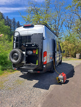 Silver RV rental van with a spare tire and utility rack, connected to a red Honda generator for power.