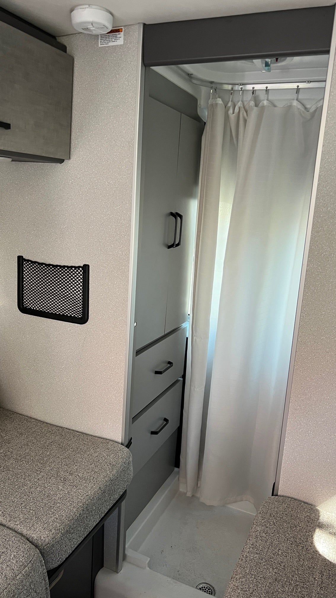 This RV rental interior showcases a compact shower with curtain, functional gray cabinets, bench seating, and a practical mesh storage net.