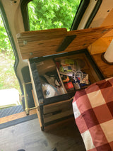 RV rental interior: An open wooden storage unit filled with various items, a brass handle, and red/white checkered bedding. Green foliage is visible through the window.