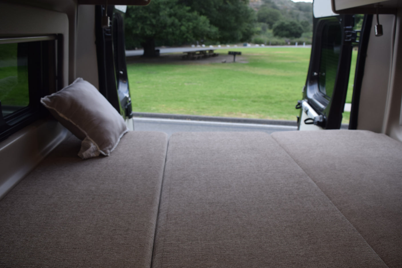 Comfy RV rental bed overlooks a green park, inviting travel and adventure from inside the camper van.