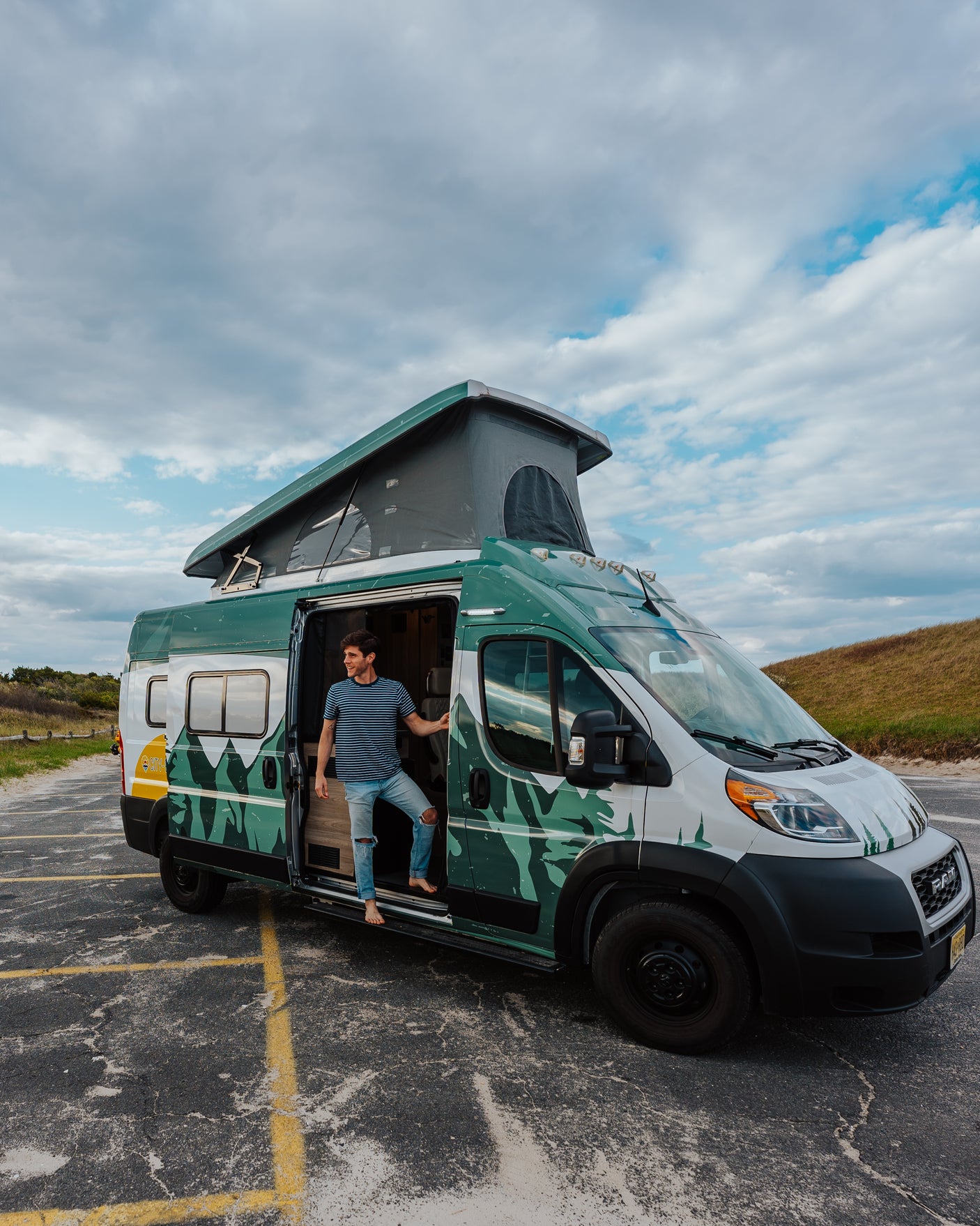 Enjoy freedom with an RV rental! A man exits a custom green & white pop-top camper van on an asphalt lot near dunes.
