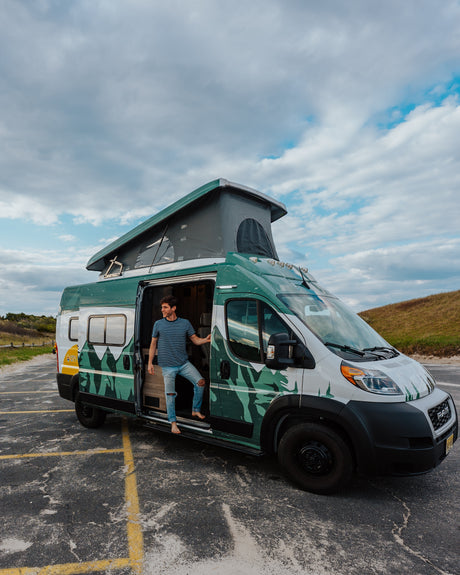 Enjoy freedom with an RV rental! A man exits a custom green & white pop-top camper van on an asphalt lot near dunes.