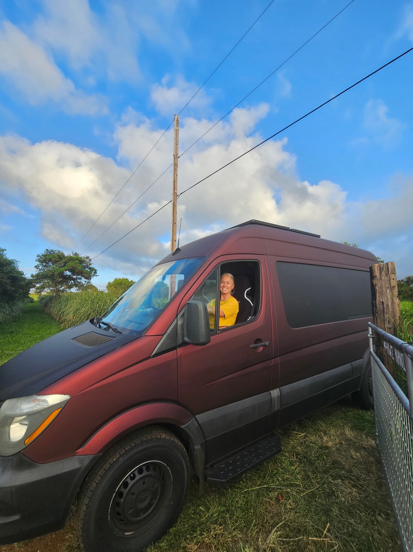 Smiling woman in a maroon rv rental van, parked in a grassy area under a bright blue, cloudy sky.