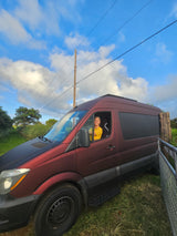 Smiling woman in a maroon rv rental van, parked in a grassy area under a bright blue, cloudy sky.