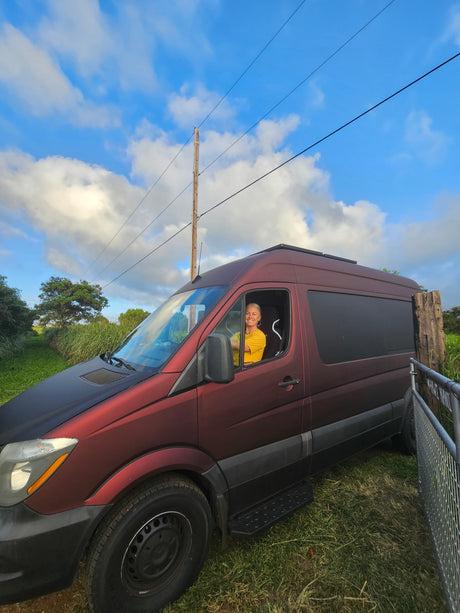 Smiling woman in a maroon rv rental van, parked in a grassy area under a bright blue, cloudy sky.