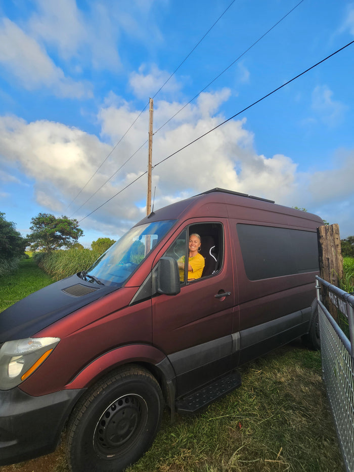 Smiling woman in a maroon rv rental van, parked in a grassy area under a bright blue, cloudy sky.