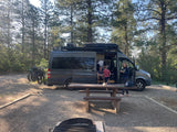 Grey RV rental van at a sunny campsite with family, bikes, picnic table, & fire pit in a pine forest.