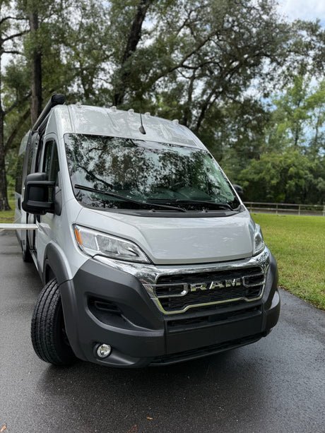 A gray Ram ProMaster camper van, wet from rain, parked on a paved surface surrounded by trees. Excellent RV rental choice.