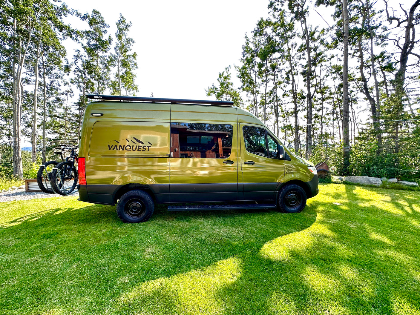 This green Vanquest camper van with bikes, parked on a lush lawn among tall trees, is ready for your rv rental adventure.