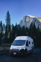White rv rental camper van parked in Yosemite, with the towering Half Dome and tall pine trees under a clear blue sky.