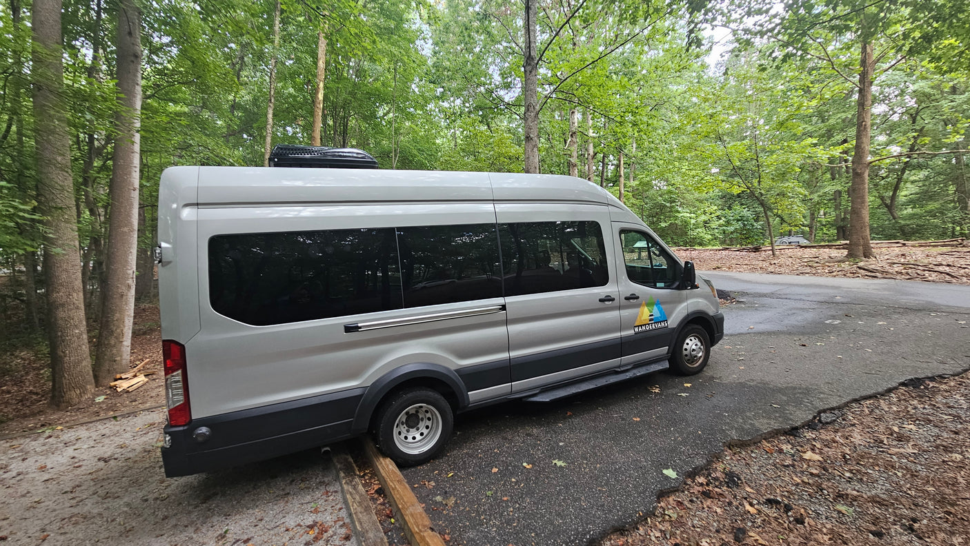 This silver Wandervans camper van, an **rv rental**, is parked on a paved road in a dense forest. A Dometic AC unit is on its roof.