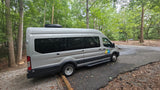 This silver Wandervans camper van, an **rv rental**, is parked on a paved road in a dense forest. A Dometic AC unit is on its roof.