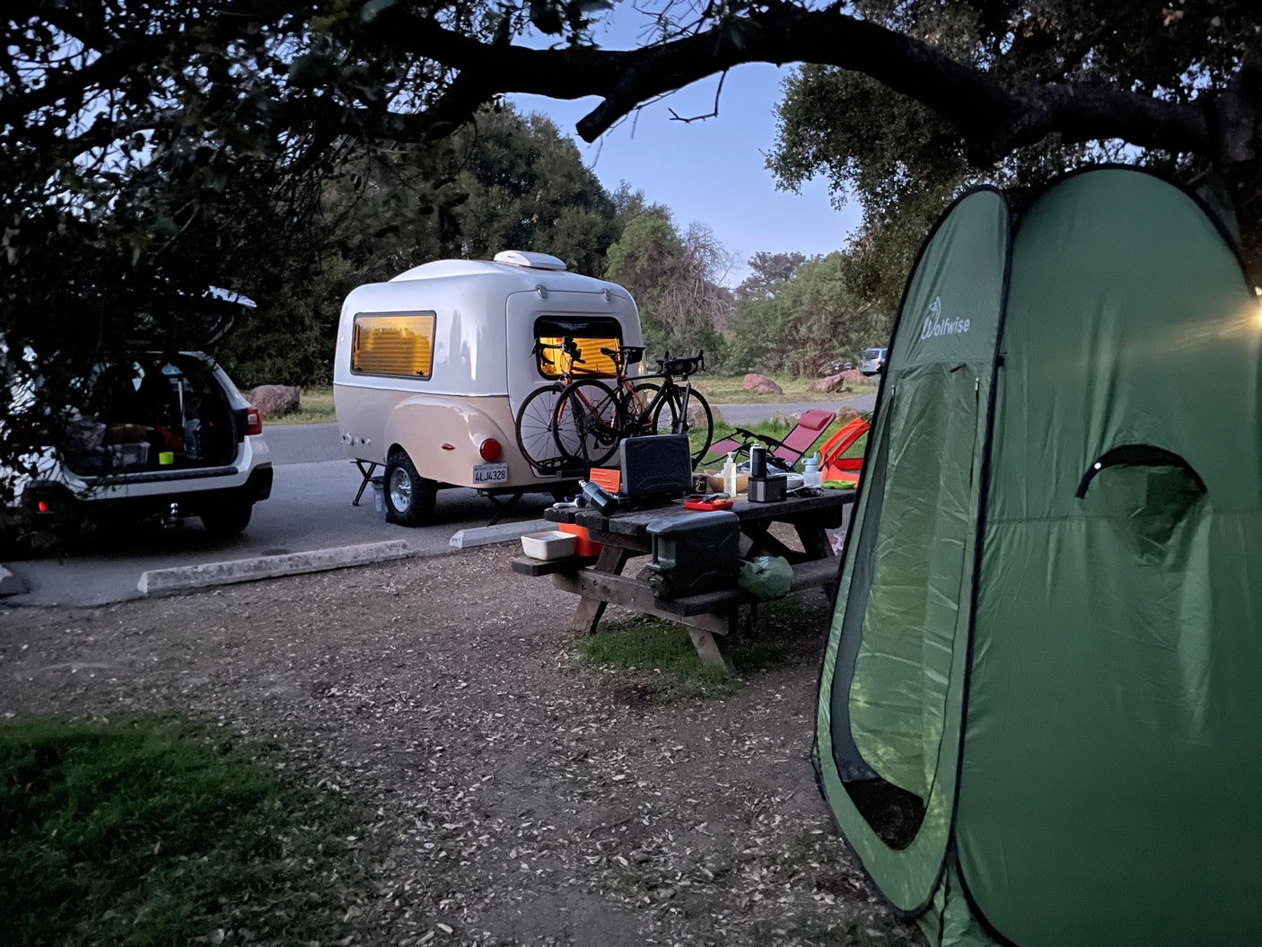 Campsite at dusk featuring an RV rental teardrop camper, bikes, car, picnic table setup, and a green tent.