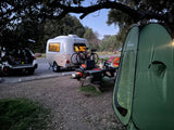 Campsite at dusk featuring an RV rental teardrop camper, bikes, car, picnic table setup, and a green tent.