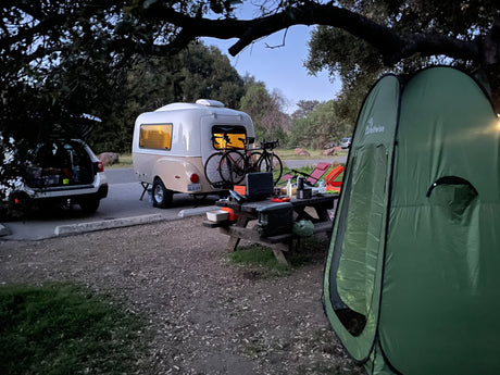 Campsite at dusk featuring an RV rental teardrop camper, bikes, car, picnic table setup, and a green tent.