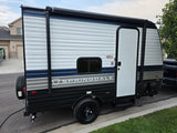 A new, compact travel trailer with blue/grey stripes and an awning, ideal for an rv rental, is parked curbside in a residential area.