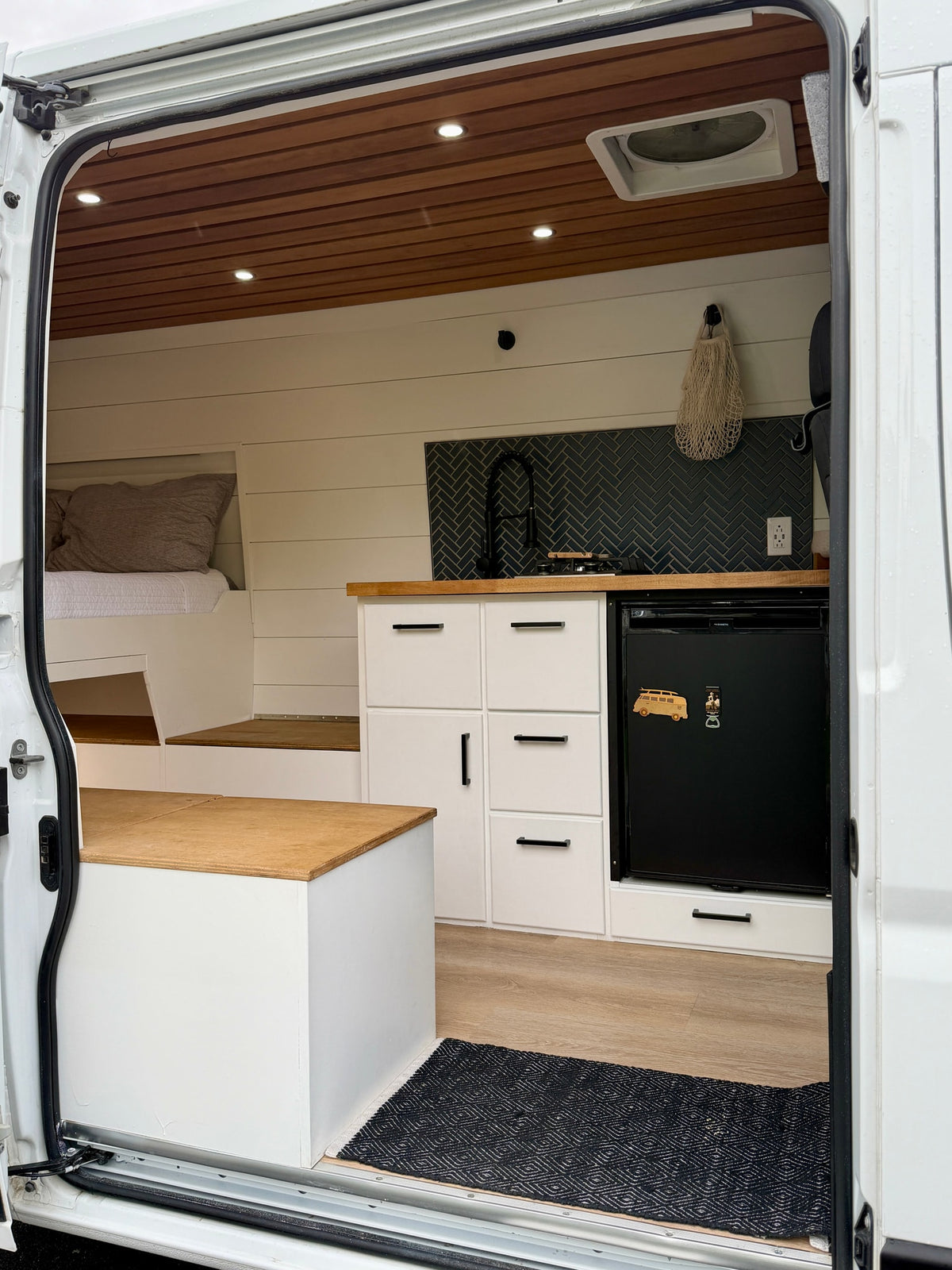 Modern RV rental interior from open side door, featuring a compact kitchen with herringbone backsplash, bed, and wood accents. — photo 5