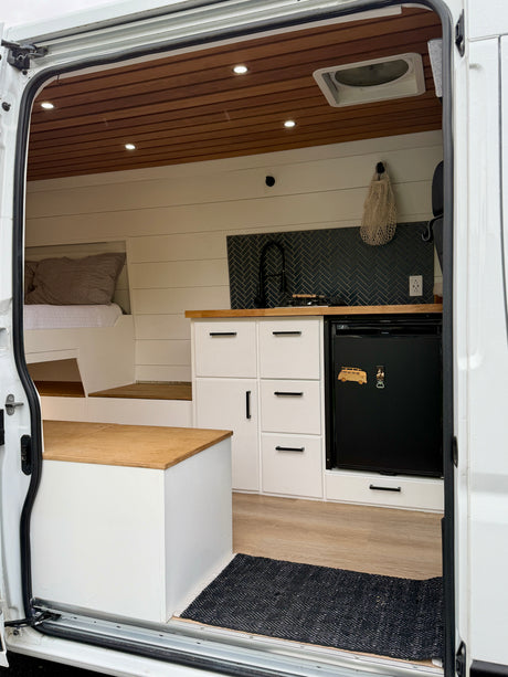 Modern RV rental interior from open side door, featuring a compact kitchen with herringbone backsplash, bed, and wood accents.