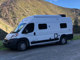 This white Winnebago Solis RV rental is parked on a gravel road, with a large, green hillside as a scenic backdrop.