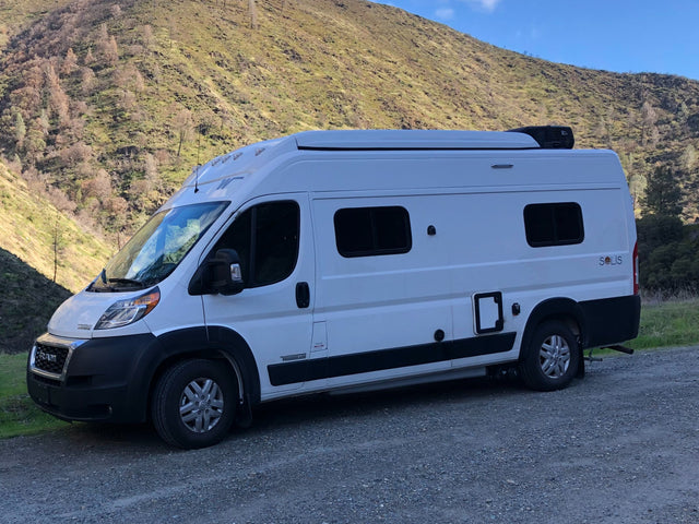 This white Winnebago Solis RV rental is parked on a gravel road, with a large, green hillside as a scenic backdrop.