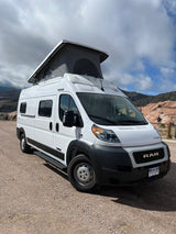 This white pop-top camper van, an RV rental, is parked on a gravel path against rocky hills and a cloudy sky.