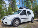 White Ford Transit Connect van with rooftop cargo box, parked in a forest campsite. Ideal RV rental for outdoor adventures.