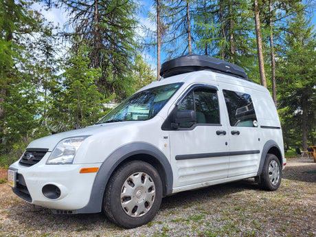 White Ford Transit Connect van with rooftop cargo box, parked in a forest campsite. Ideal RV rental for outdoor adventures.