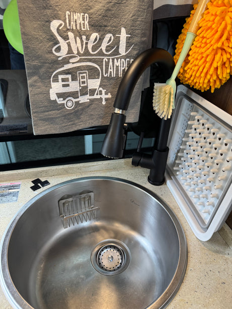 RV rental kitchen: black gooseneck faucet, stainless steel sink with dish rack, cleaning brush, and 'Camper Sweet Camper' towel.