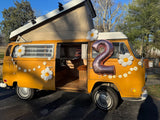 Experience a vintage yellow VW van RV rental! This pop-top camper, adorned with daisies and a big '2' balloon, is ready for fun.