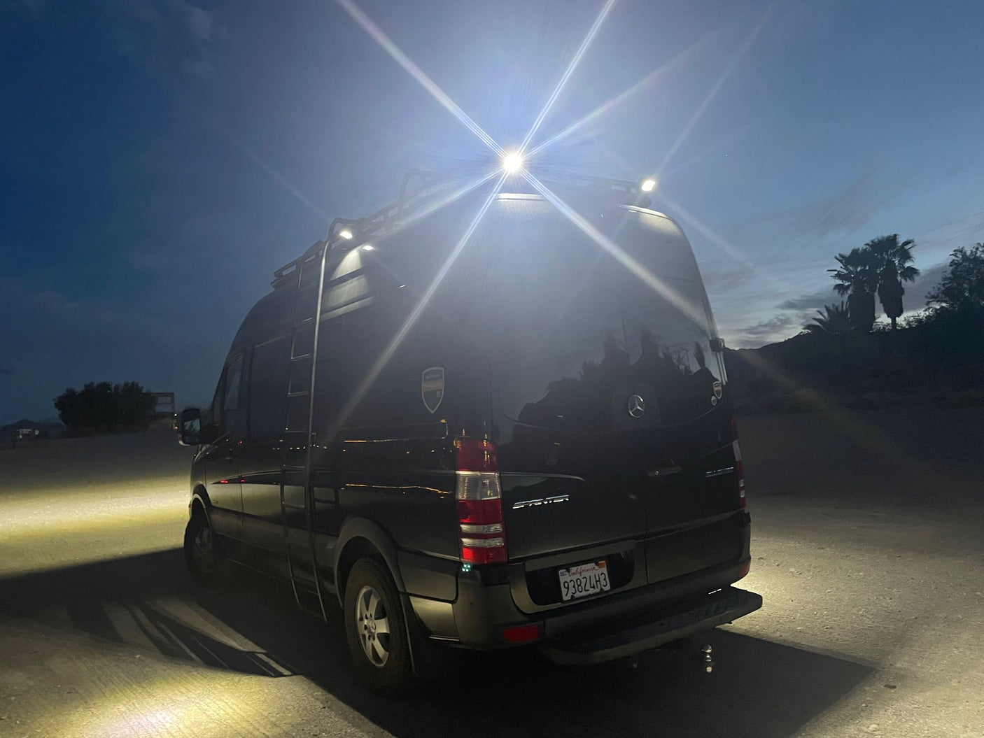 Black Mercedes Sprinter camper van rv rental, bright roof lights on, parked on gravel at night.