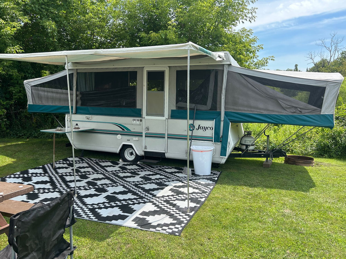 White Jayco pop-up RV rental fully set up at a green campsite with an extended awning and a patterned outdoor mat.