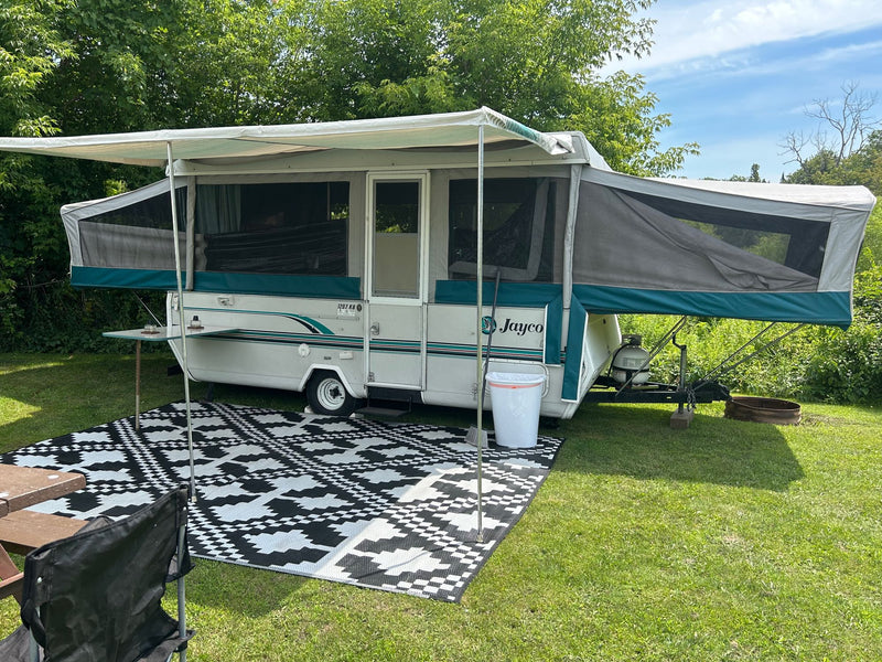 White Jayco pop-up RV rental fully set up at a green campsite with an extended awning and a patterned outdoor mat. — photo 1
