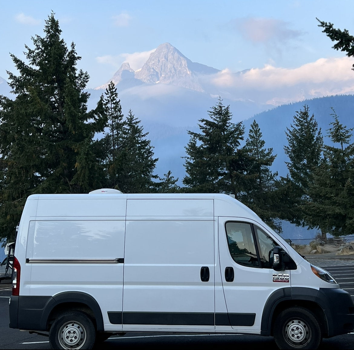 White RV rental van at a scenic mountain viewpoint with evergreen trees and majestic peaks.|Cozy rv rental camper van interior: comfortable bed