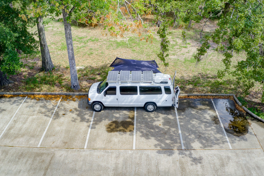Aerial shot: White rv rental van with roof solar panels and side awning parked in a lot under trees for off-grid camping.