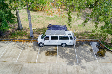 Aerial shot: White rv rental van with roof solar panels and side awning parked in a lot under trees for off-grid camping.