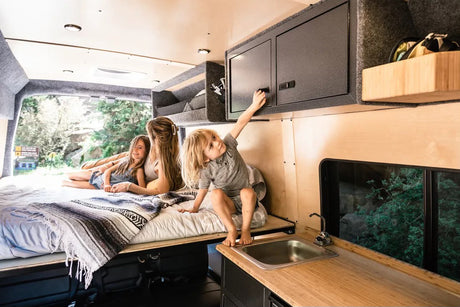 Family fun in an rv rental: A mother and two kids relax on the bed, a child reaches for overhead cabinets, a sink is visible.