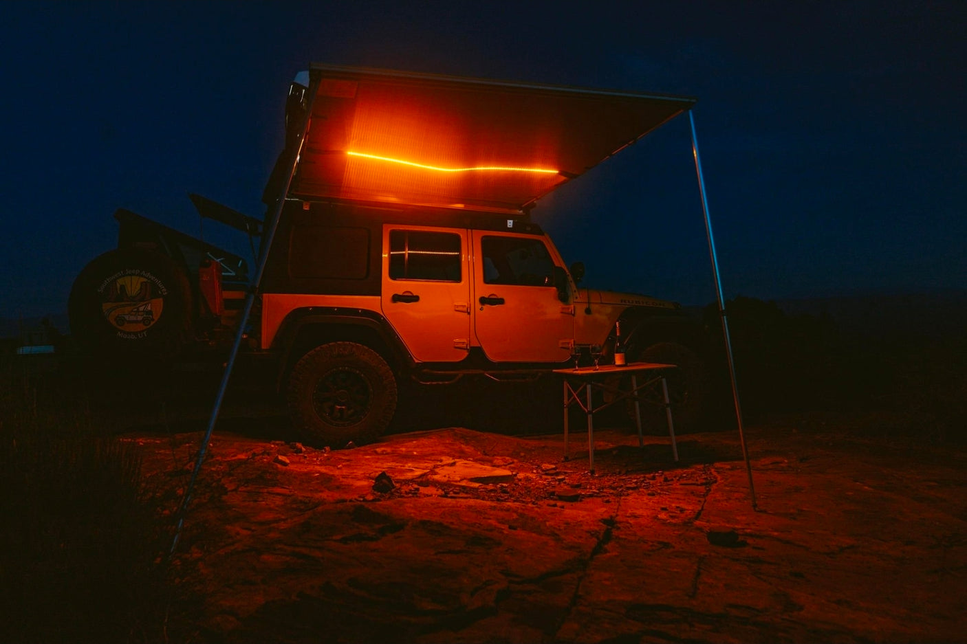 Experience night overlanding with this RV rental Jeep. Illuminated awning, table with drinks, perfect for a desert adventure.