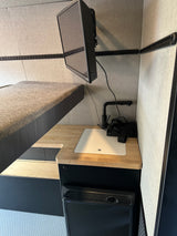 Modern RV rental interior with bunk bed, wall-mounted TV, wood counter, sink, & Engel fridge. Compact living.