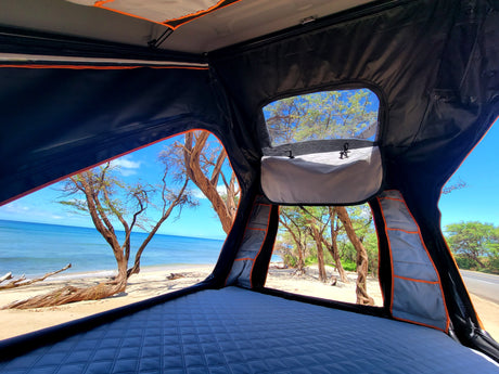 Serene ocean view from an RV rental rooftop tent. Interior shows a comfy mattress, framed by trees and blue sky.