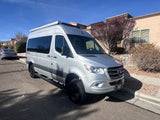 A silver Mercedes-Benz Sprinter camper van for RV rental, parked on a sunny residential street.